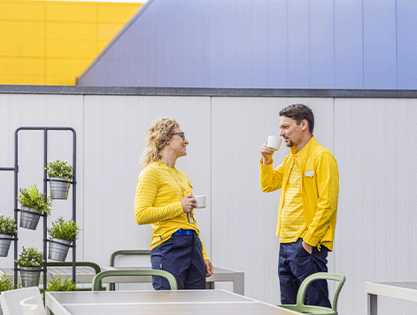 Two store employees in yellow and blue uniforms standing on an outdoor patio, chatting and smiling while drinking coffee from white mugs.
