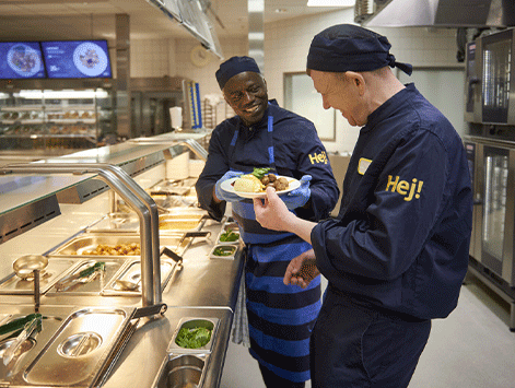 Two smiling kitchen employees in dark blue uniforms with "Hej!" on the sleeves share a plate of food over a commercial cafeteria service counter.