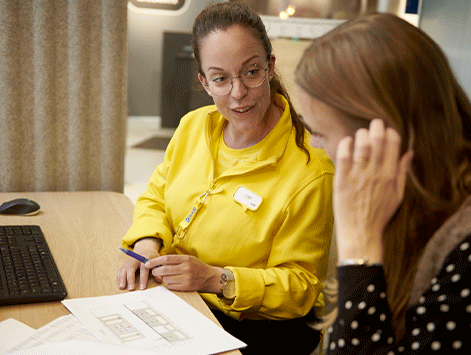 A retail employee in a bright yellow uniform jacket and glasses points at a printed floor plan design while speaking with a customer at a wooden desk.