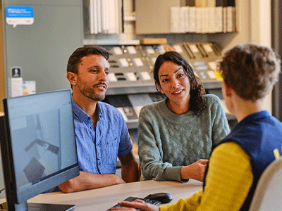 A couple sitting at a desk and smiling while speaking with an IKEA employee who is working on a 3D kitchen design layout on a computer screen.