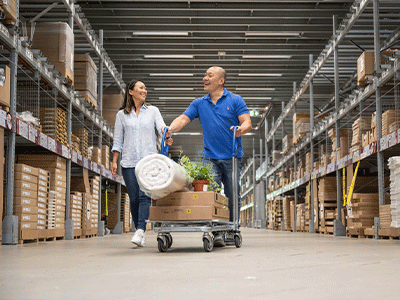 A happy couple smiling and walking down a warehouse aisle while pushing a flatbed trolley loaded with flat-packed boxes, a rolled rug, and a plant.