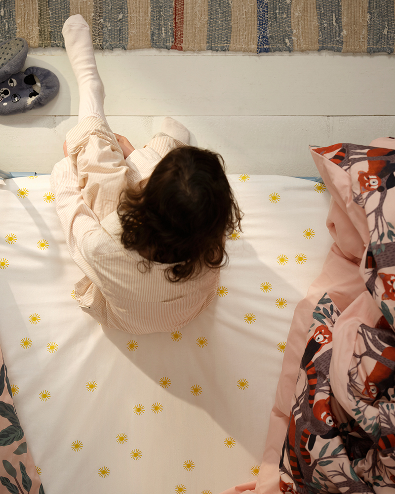 A girl sitting on a bed with yellow-speckled sheets, surrounded by cushions featuring red pandas from the ALPTALL collection.