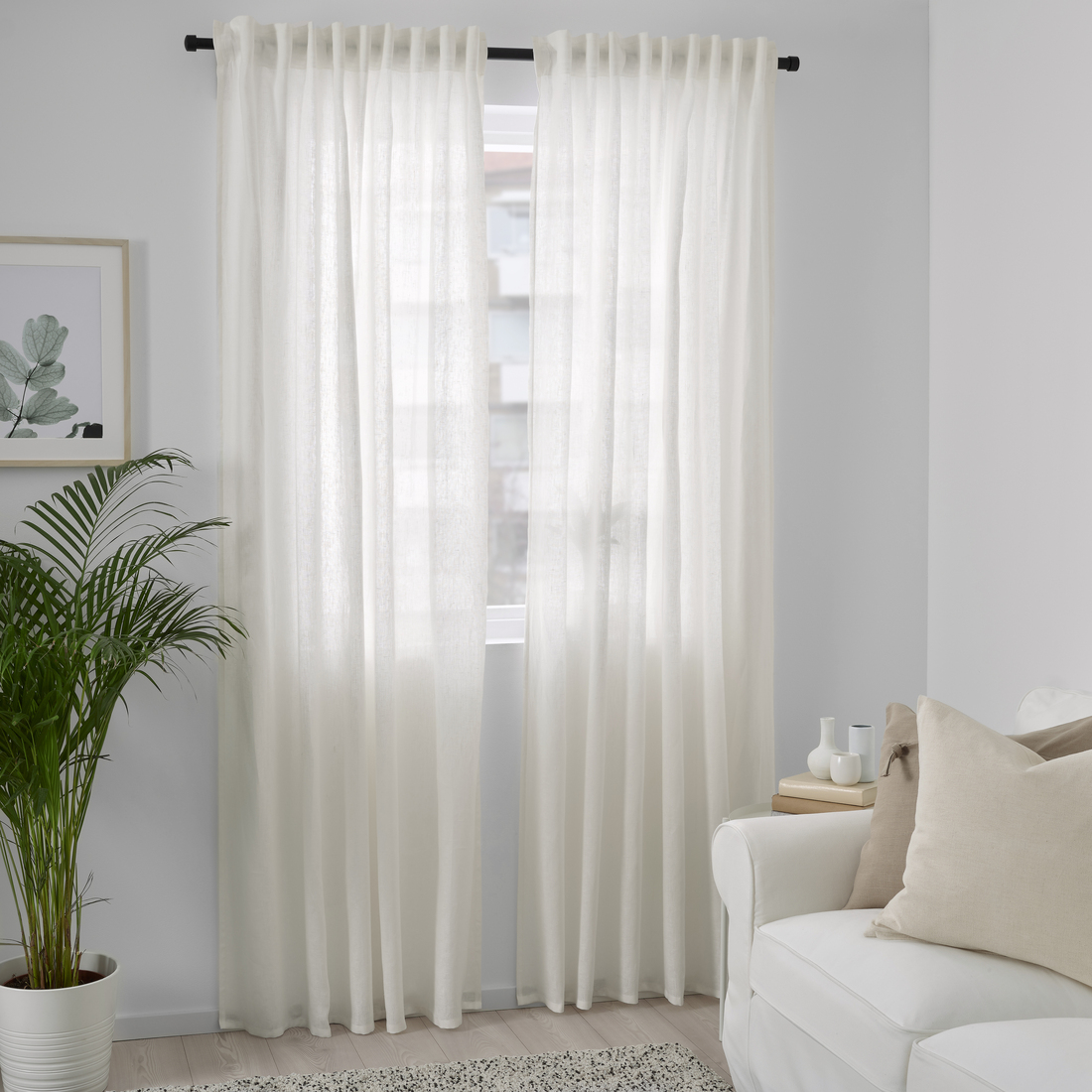 Cortinas traslúcidas de color blanco colgadas en una barra negra frente a una ventana, en una sala con un sofá blanco y una planta de interior.