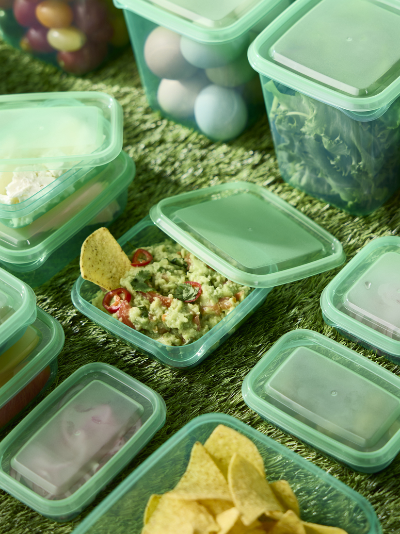 Vista superior de múltiples recipientes herméticos de plástico verde sobre césped artificial, uno de ellos con guacamole y totopos.