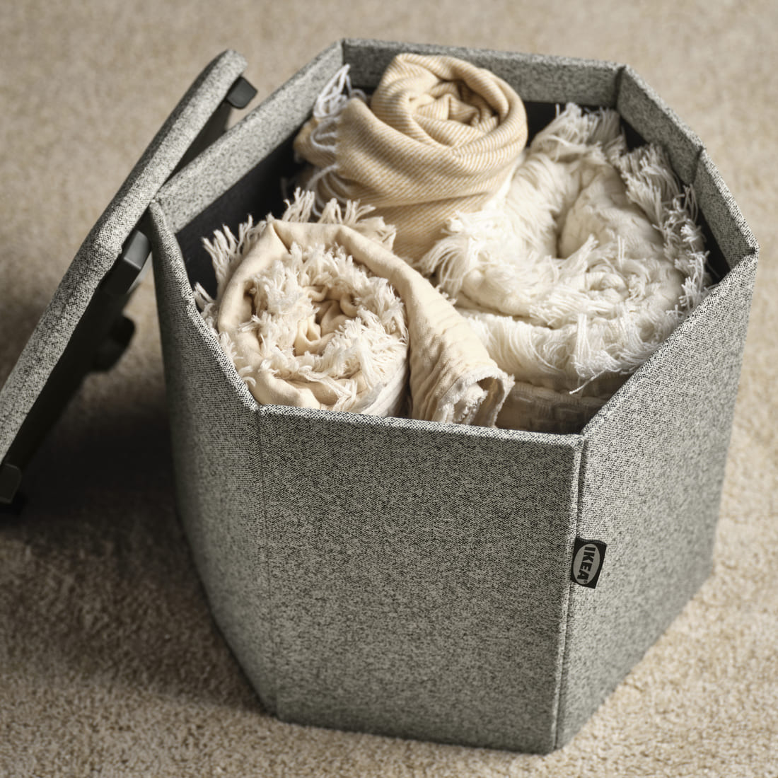 Caja de almacenaje hexagonal de tela gris con la tapa entreabierta, llena de mantas y textiles enrollados sobre una alfombra beige.