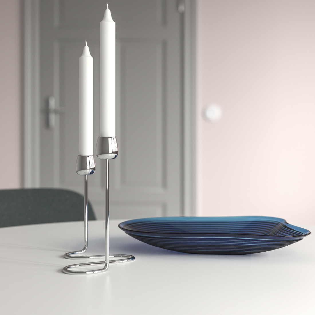 Dos velas blancas sobre candelabros metálicos cromados de diseño ondulado junto a un plato de vidrio azul sobre una mesa blanca.