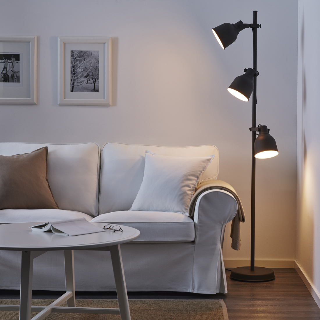 Lámpara de pie metálica negra con tres focos orientables en una sala iluminada junto a un sofá blanco y una mesa de centro.