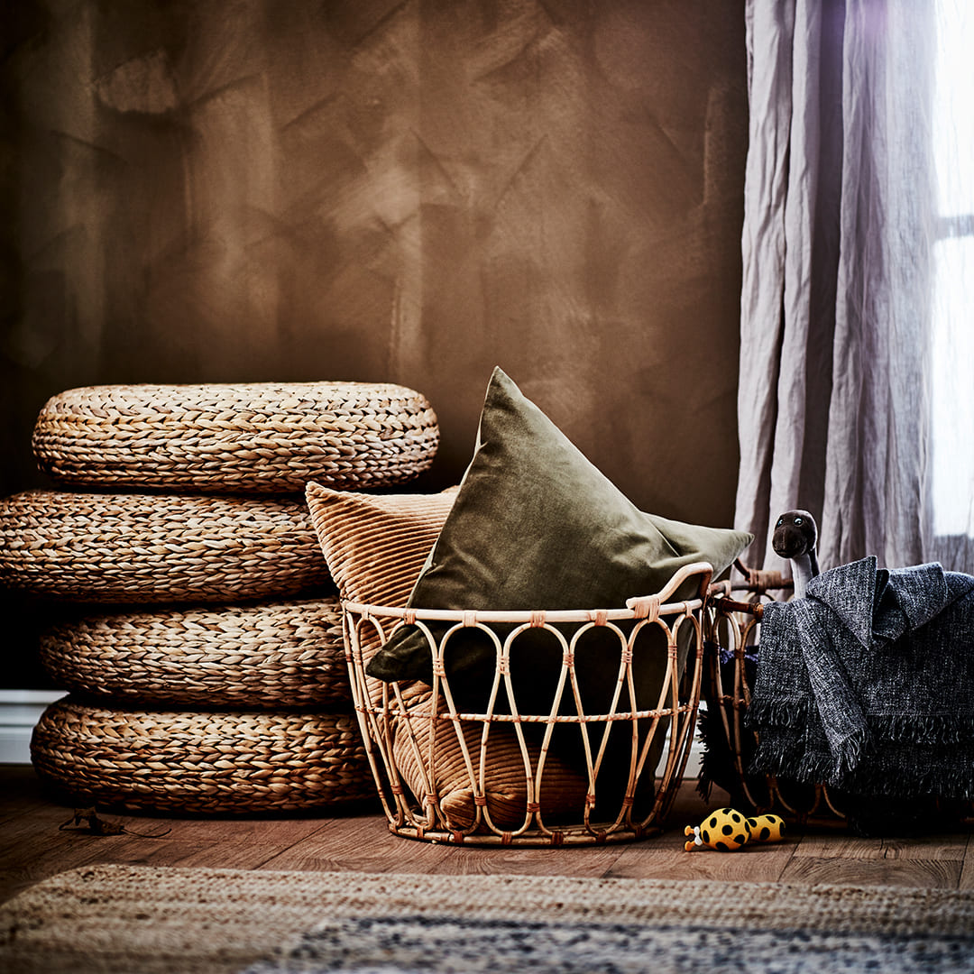 Rincón decorativo de estilo rústico con cojines apilados de fibra natural, una cesta de mimbre con cojines de felpa y mantas cerca de una ventana.