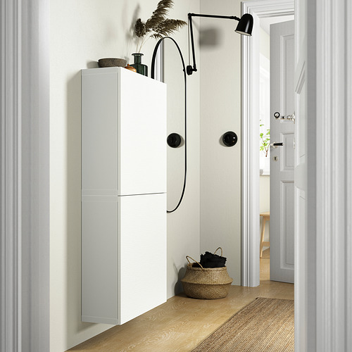 Mueble de almacenaje BESTÅ blanco con dos puertas. Montado en la pared, complementado con cesta tejida y lámpara de pared. Diseño alto y estrecho.