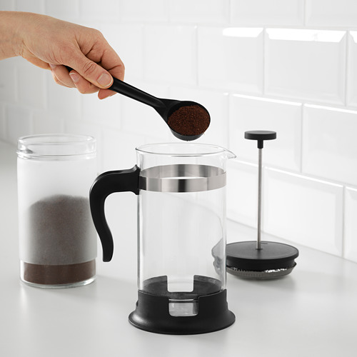 Una mano vierte los posos de café en una prensa francesa de vidrio transparente, con un émbolo listo para preparar en una encimera blanca.