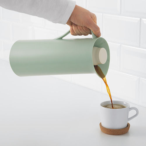 La persona vierte café de un matraz verde al vacío en una taza blanca en un posavasos de corcho.