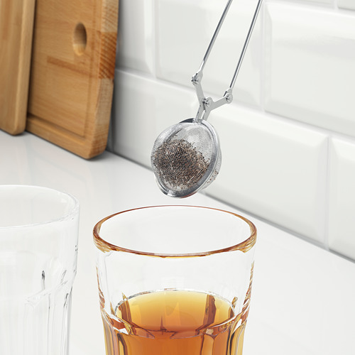 El infusor de té de plata sostenido por pinzas se sumerge en un vaso transparente de té dorado junto a una jarra a juego.