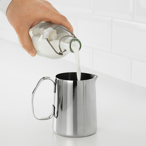 Una mano vierte la leche de un biberón en una jarra espumadora de leche MÅTTLIG de acero inoxidable, mostrando su diseño duradero y fácil de limpiar.
