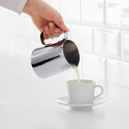 Verter a mano la jarra de leche espumosa en una taza de café blanco en el platillo, demostrando la espuma de la leche.