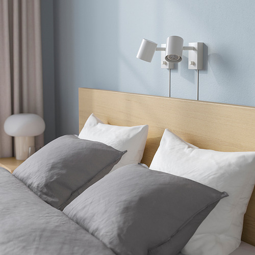 Cama moderna con cabecero de madera, dos apliques regulables encima, almohadas y manta grises, pared celeste.