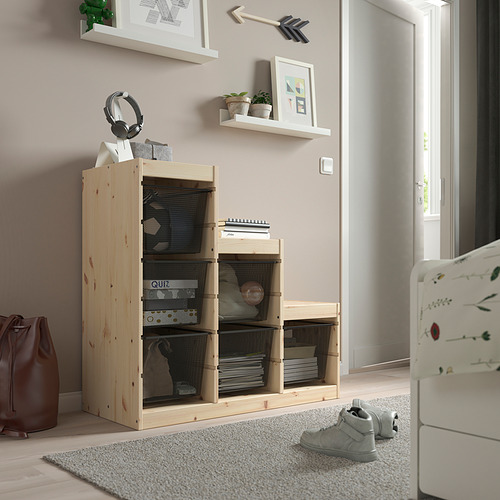 Mueble de almacenamiento TROFAST de madera de color claro con cajas de malla, que contienen varios artículos, colocados cerca de una puerta y muebles blancos.