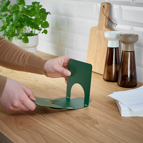 Persona montando servilletero de metal verde sobre mesa de madera, cerca de plantas y condimentos.