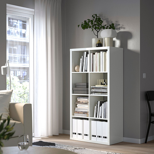 Estantería KALLAX blanca con libros y decoración, junto a ventana, en salón gris.