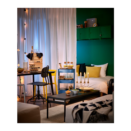 Acogedor dormitorio con paredes verdes, mesa de centro negra, silla amarilla, cortinas transparentes, carrito azul, armario verde sobre cama con almohada amarilla.