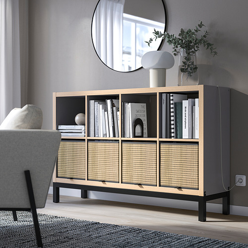 Salón moderno con estantería KALLAX, madera clara y negro. Exhibiendo libros y decoración. Espejo arriba, silla gris al lado, que se asemeja al tamaño de la habitación.