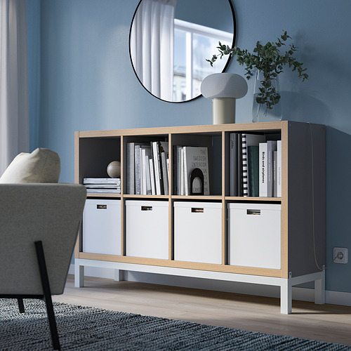 Estantería KALLAX en madera clara y blanco con libros y lámpara.