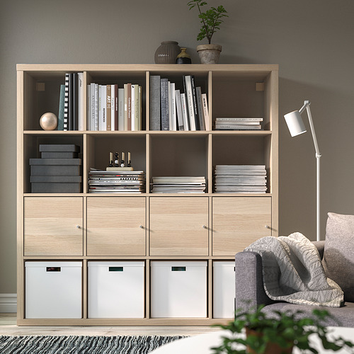 Estantería KALLAX, roble claro, forma cúbica, diseño moderno, alto, contiene muchos libros y decoración. Un acogedor sofá gris descansa al lado.