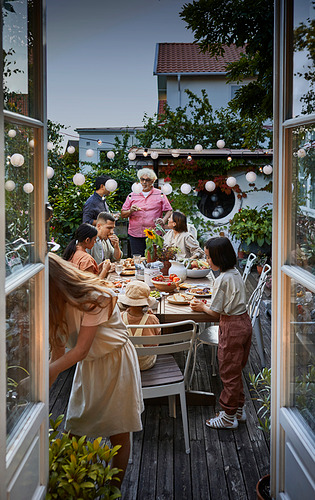 La familia disfruta de una comida al aire libre en una robusta mesa y sillas NORRMANSÖ de madera, rodeadas de luces y vegetación.