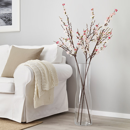 SMYCKA: jarrón alto y transparente con flores de color blanco rosado junto a un sofá blanco, con un diseño natural pero artificial.