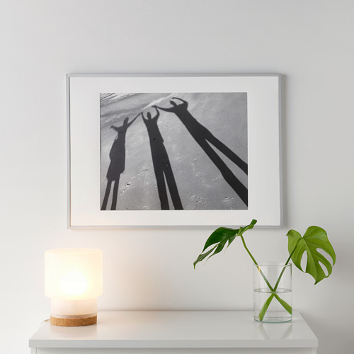A white picture frame a black-and-white shadows on a white shelf. Beside it, a small lit lamp and a glass vase with a green plant.