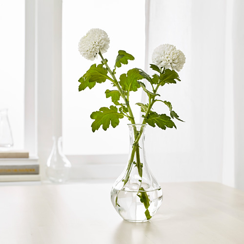 Un pequeño jarrón de vidrio, VILJESTARK, contiene dos flores blancas con tallos y hojas verdes. Se muestra sobre una superficie blanca.