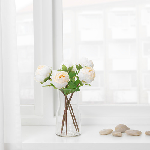 Un jarrón de vidrio transparente con un soporte de cruz con tallo muestra flores blancas, melocotón y hojas verdes. Se asienta sobre una superficie blanca cerca de una ventana.