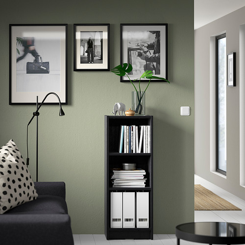 Estantería negra BILLY con libros y decoración, arte enmarcado, lámpara, sofá y almohada en una habitación verde con azulejos blancos.