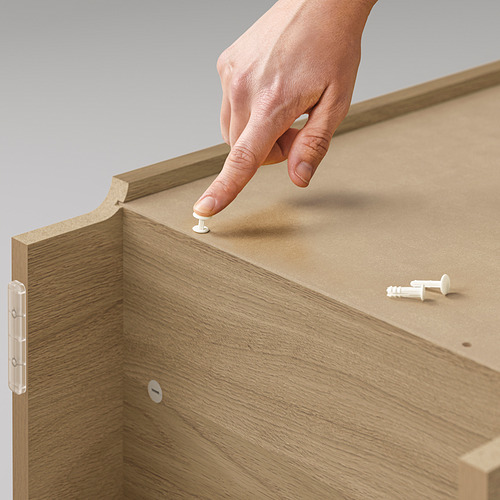 Una mano instala una bisagra blanca en el lateral de una estantería de madera beige BILLY.