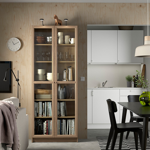 Sala de estar moderna con librería BILLY. Exhibiendo platos, libros y decoración. Al lado una cocina blanca con sillas negras.