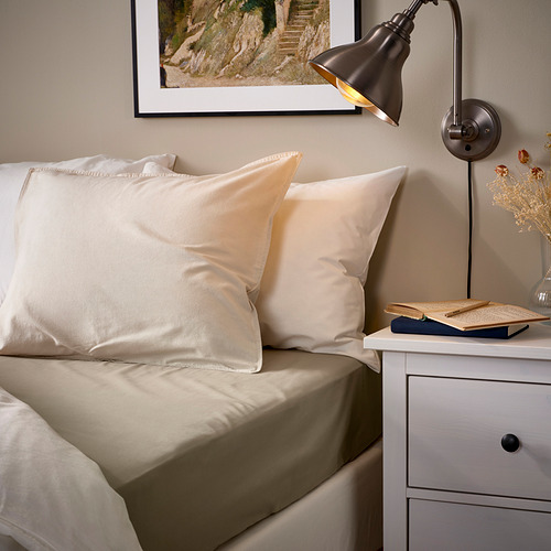 Acogedora cama con sábanas beige ULLVIDE, lámpara negra, cuadro, libros, jarrón. Las sábanas se ajustan al colchón grueso, la lámpara de pared proporciona luz.