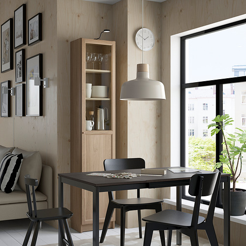 Comedor moderno con mesa negra, estantería BILLY, lámpara colgante, sofá acogedor y ventana con vista a la vegetación.