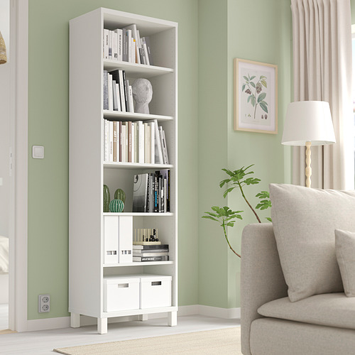 Librería blanca BESTÅ contra la pared verde, que contiene libros y decoración. Sillón beige, lámpara de pie y planta cerca.