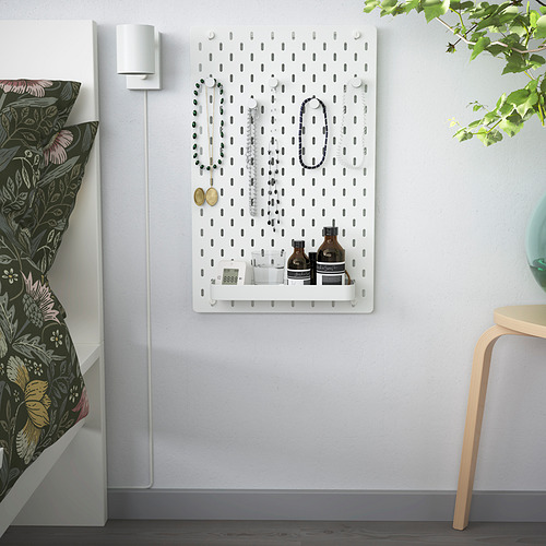 Tablero perforado SKÅDIS con joyas, botellas, reloj. Al lado de la cama con almohadas florales y silla.