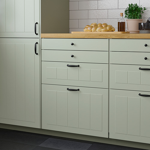 Cocina moderna con mueble verde claro, manijas oscuras, encimera de madera. Pan, botellas y plantas expuestas.