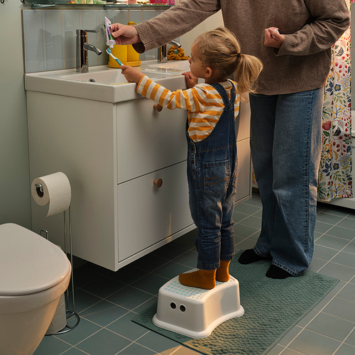 Un niño está de pie en un taburete blanco FÖRSIKTIG en un baño, cepillándose los dientes con la ayuda de un adulto. Suelo de baldosas verdes, lavabo blanco.