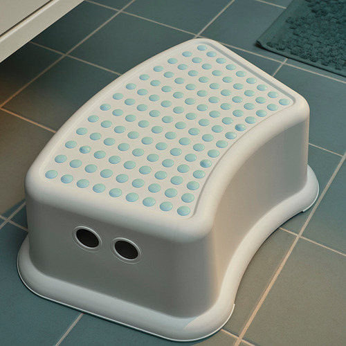 Taburete blanco para niños con textura de puntos azules, bordes redondeados y parte inferior antideslizante sobre un piso de baldosas.