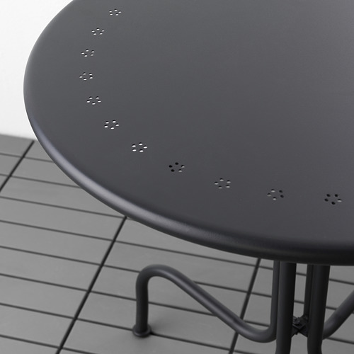 Mesa circular de exterior negra con superficie de acero duradera con recubrimiento en polvo y robustas patas metálicas, sobre suelo de baldosas.