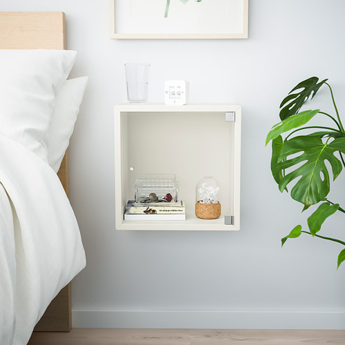 Dormitorio minimalista con estante de cubo EKET blanco para almacenamiento y exhibición, que muestra un reloj, plantas y accesorios, colocados cerca de una cama.