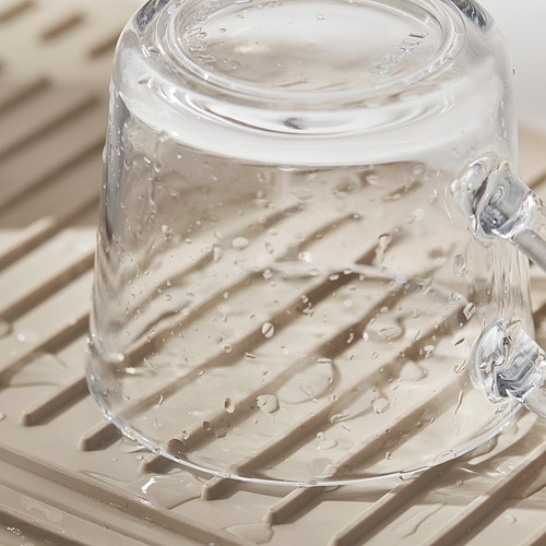 El vaso de vidrio transparente con gotas de agua se asienta sobre un tapete de color canela y texturizado, probablemente un tapete de secado expandible y con agarre.
