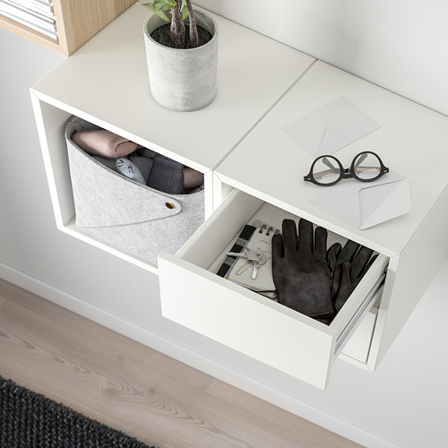 Mueble de almacenamiento de pared blanco, accesorios de sujeción. Cajón abierto, mostrando guantes, llaves y un cuaderno. La parte superior tiene una planta y ropa doblada.