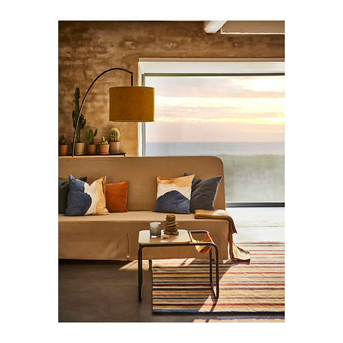 Acogedora sala de estar con sofá color canela, almohadas de colores, alfombra a rayas, lámpara y cactus cerca de grandes ventanales que muestran una puesta de sol.