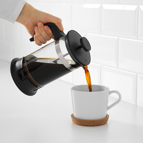 La persona vierte café recién hecho de una prensa francesa en una taza blanca en un posavasos de corcho.