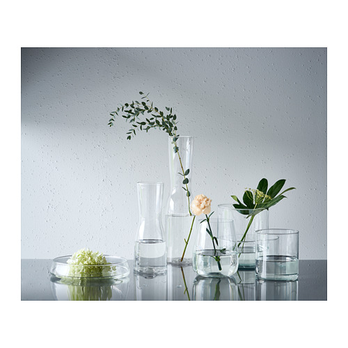 Seis jarrones de vidrio transparente de diferentes tamaños exhiben flores sobre una superficie blanca contra una pared gris.