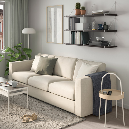 Sala de estar moderna con sofá beige, alfombra gris, mesa auxiliar, taburete, jarrones, arte, estantes con libros y plantas.