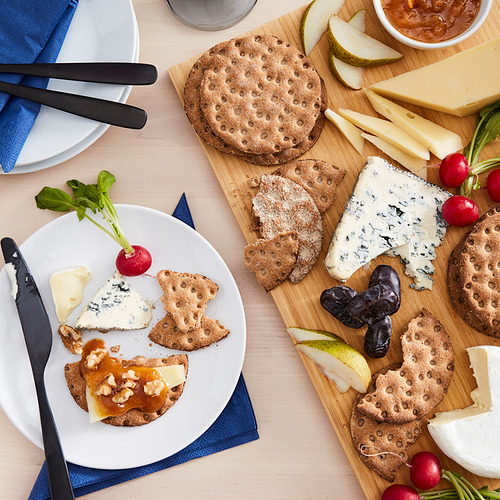Vista superior de una configuración de bocadillos con pan crujiente KNÄCKEBRÖD RÅG, quesos, galletas saladas, frutas y rábanos en una tabla y un plato de madera.